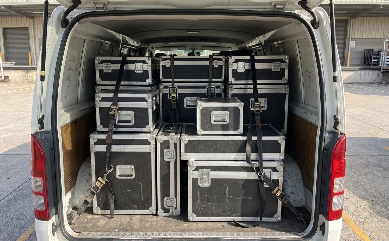 ハイエースの荷室に機材ケースが効率よく積まれている様子。ケース同士が隙間なく配置され、固定用のストラップが見える。人物なし。