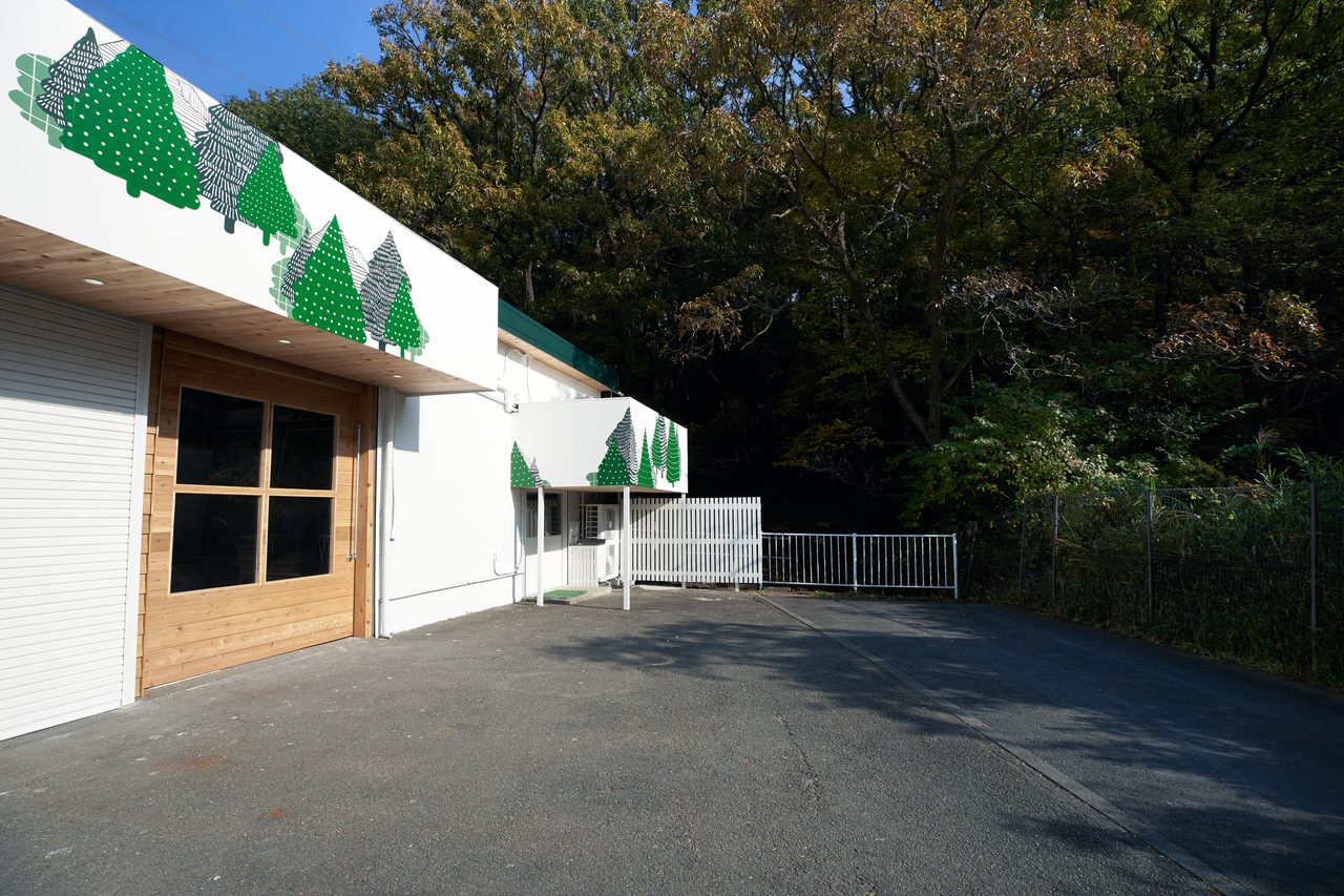 スタジオの外観。白い建物の前に広がる駐車場と、周囲を取り囲む緑豊かな木々。手前にはアスファルトの地面と少しの雑草が見える。青空の下で、自然の中に静かに佇む建物の雰囲気。
